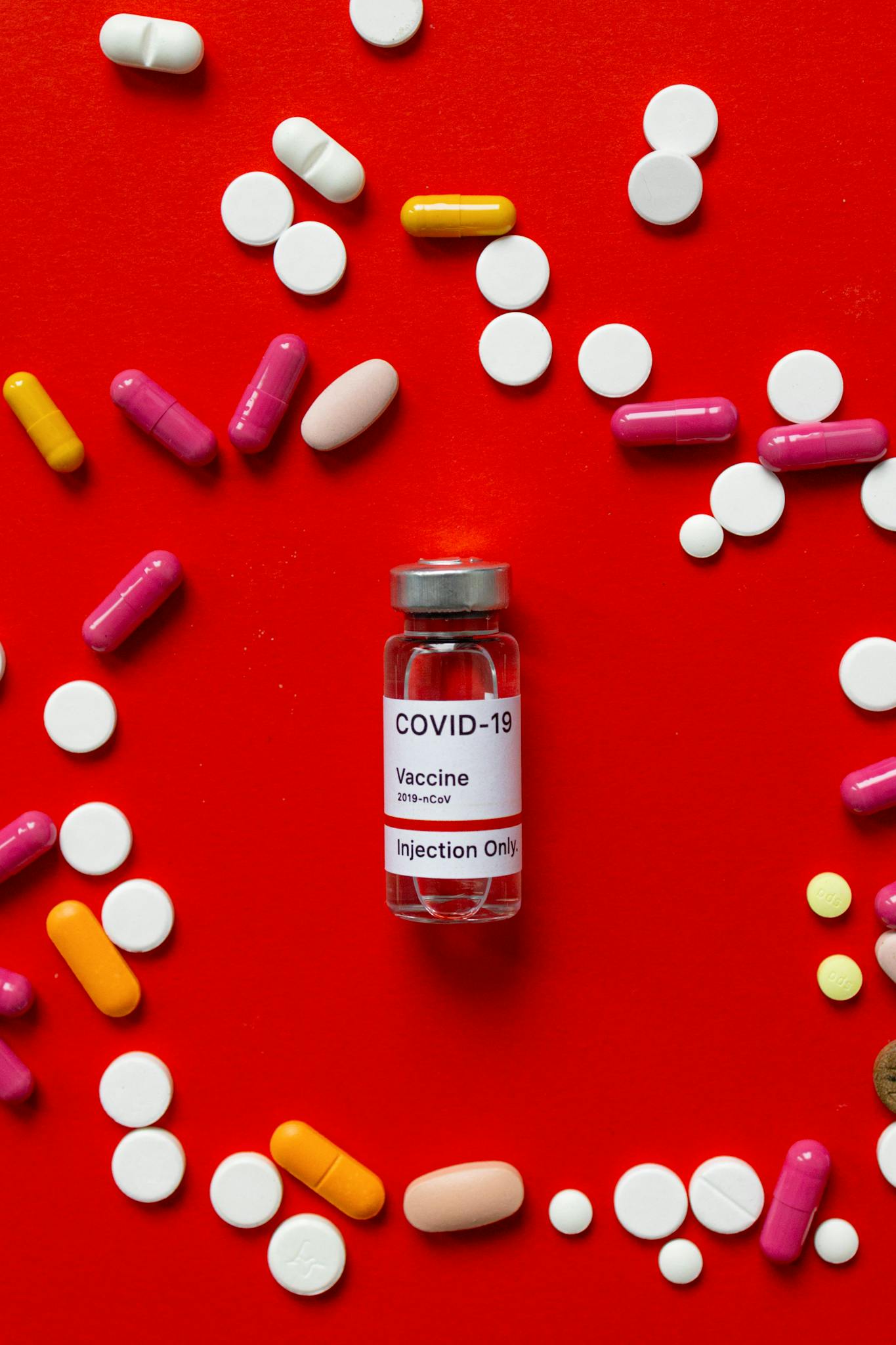 Colorful medication pills and COVID-19 vaccine vial on red backdrop, health concept.