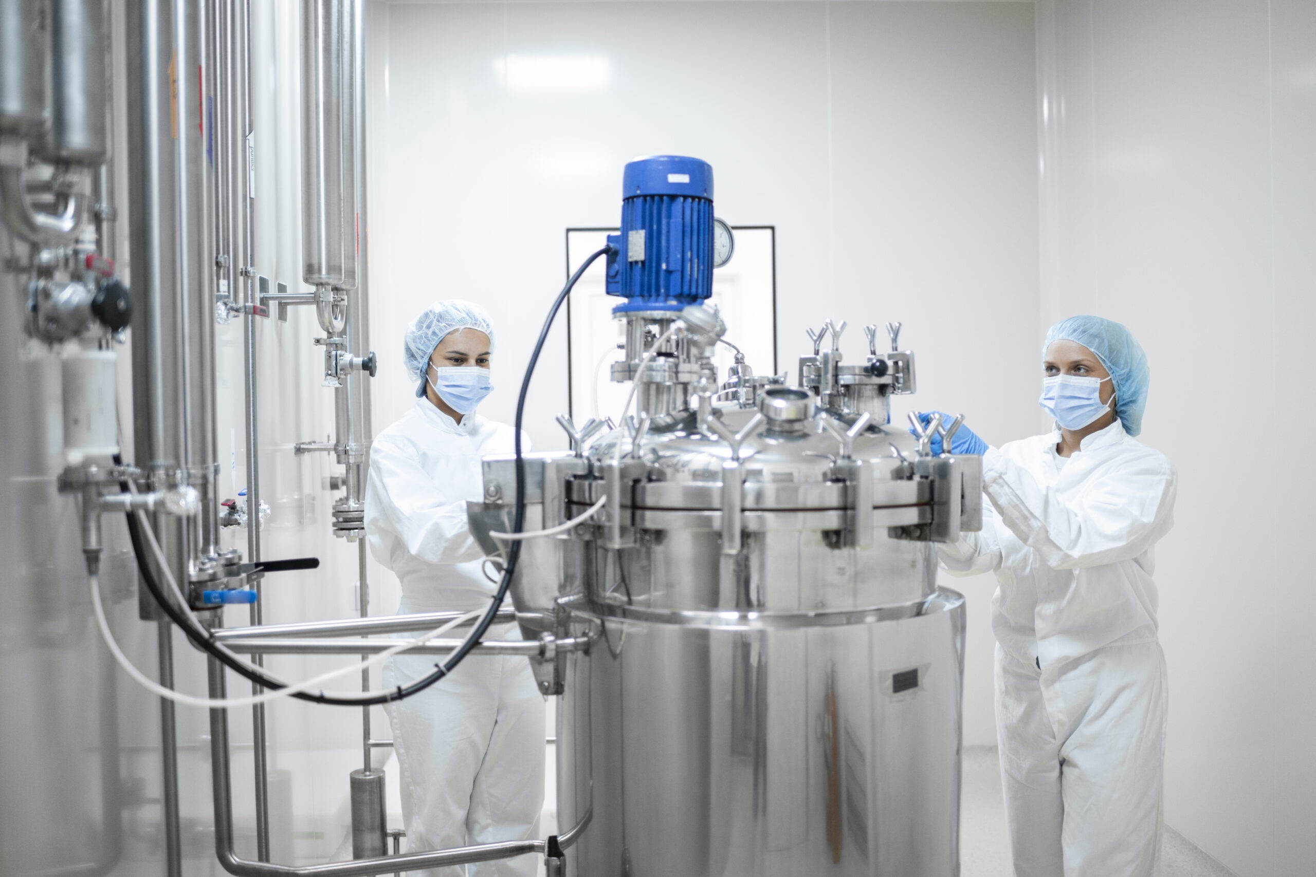 Two workers in protective gear operating stainless steel tanks in a pharmaceutical production facility.